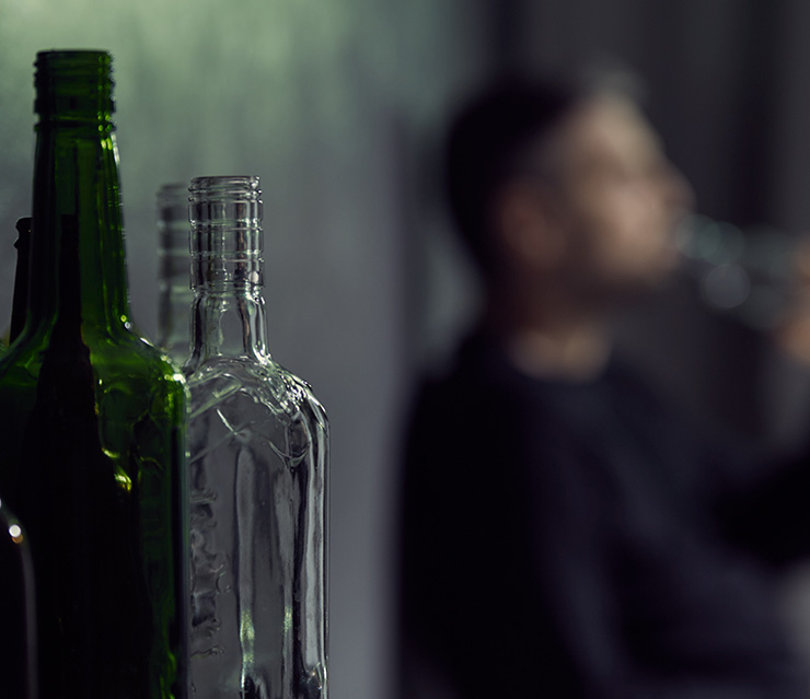 Mehrere leere Glasflaschen im Vordergrund, im unscharfen Hintergrund eine Person mit einer Flasche in der Hand in einem Innenraum.