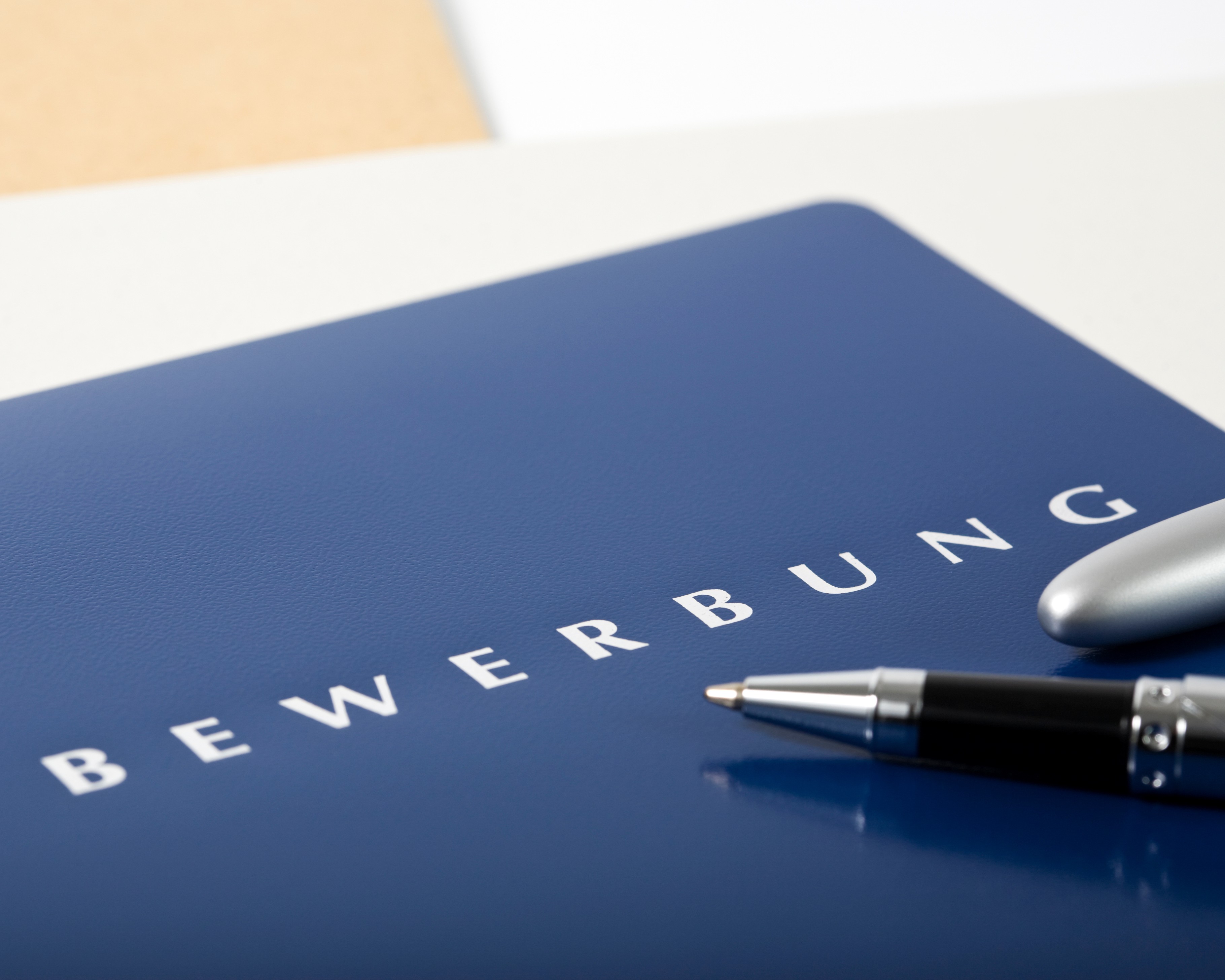 Blaue Bewerbungsmappe mit der Aufschrift „BEWERBUNG“ und einem Kugelschreiber auf einer hellen Tischoberfläche.