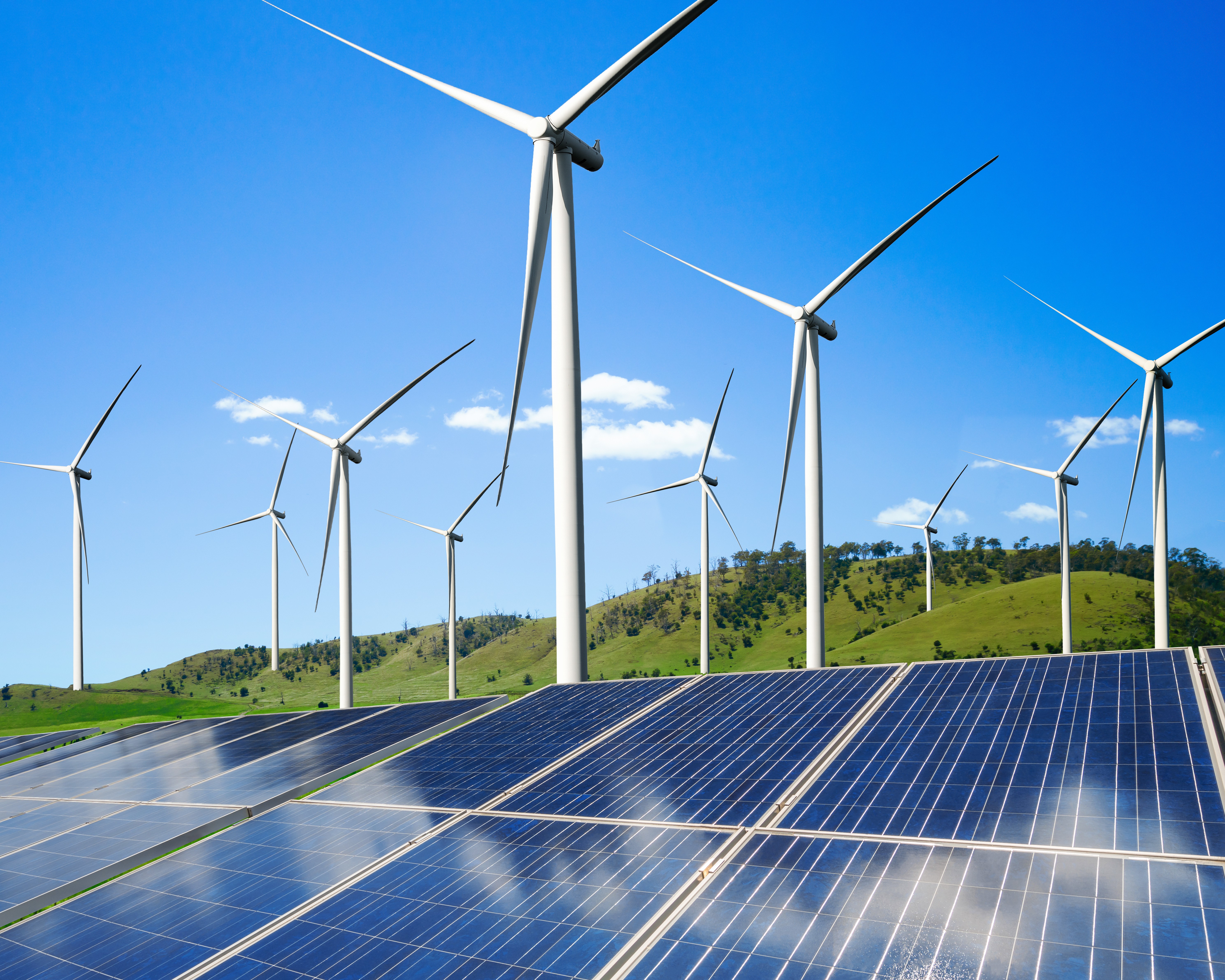 Großflächige Photovoltaikanlage vor mehreren Windkraftanlagen auf einer grünen Hügellandschaft unter blauem Himmel.