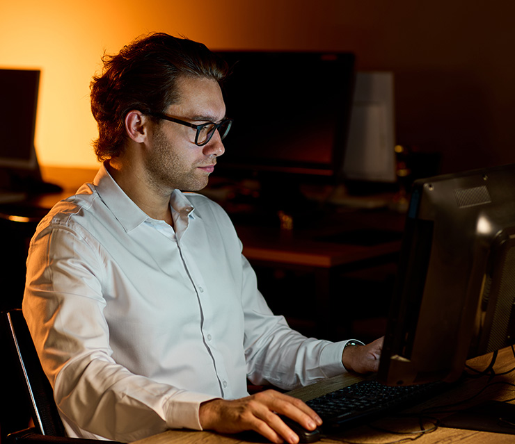 Person arbeitet abends an einem Computer in einem schwach beleuchteten Büro als Symbol für lange und überlange Arbeitszeiten.