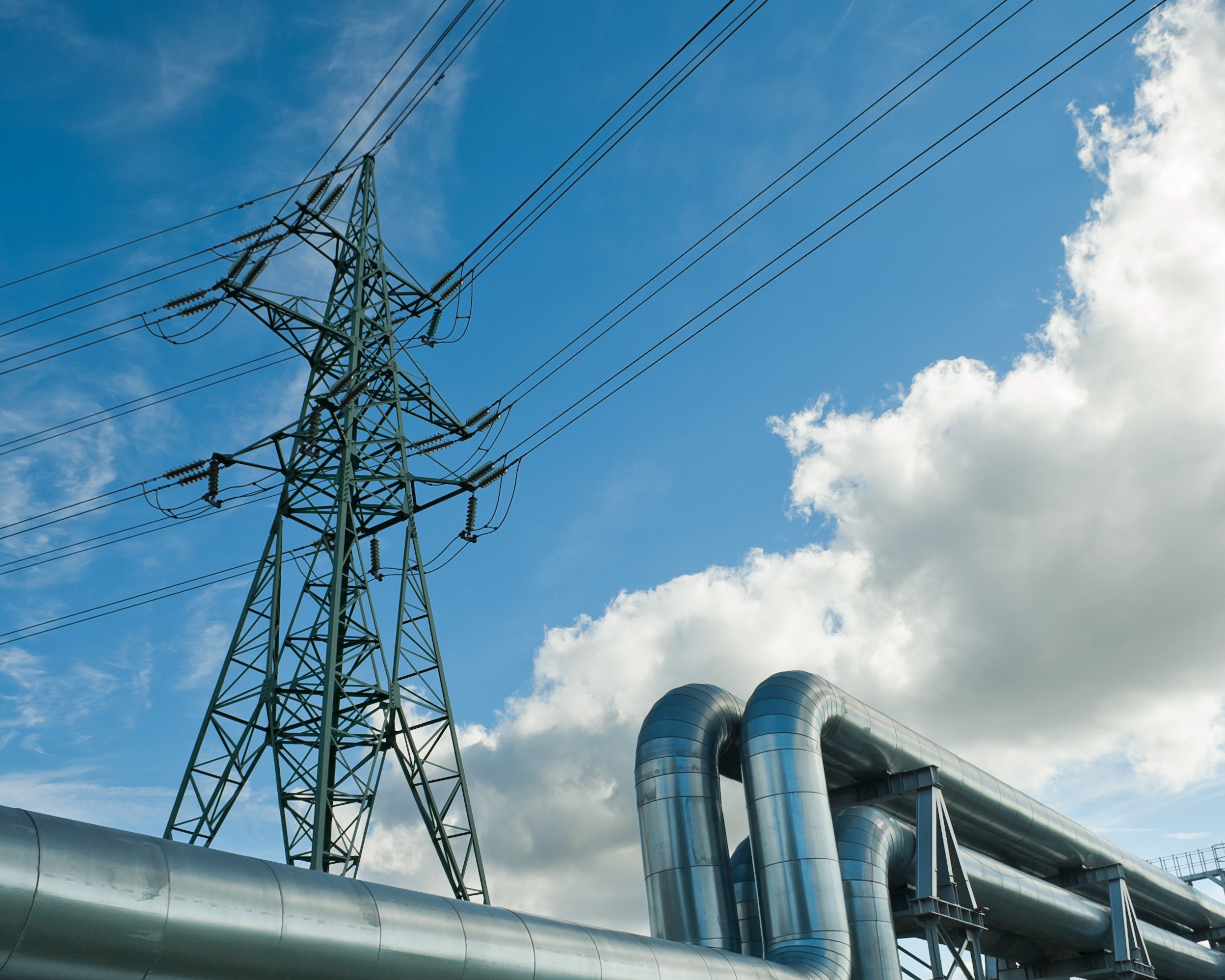 Hochspannungsmast und industrielle Rohrleitungen vor blauem Himmel mit großen Wolken auf einem Gewerbe- und Industrieareal.